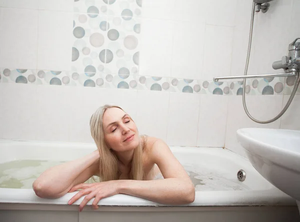 A beautiful smiling sexy girl with blond hair sits in a bath and washes, in the bathroom. An image about cleanliness and about taking care of yourself and your body.