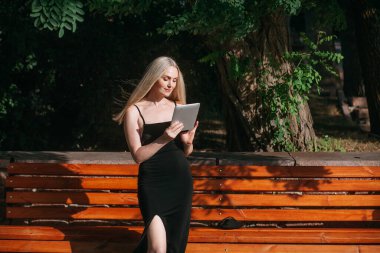 Uzun sarı saçlı, uzun siyah uzun elbiseli, pozitif bir kadın, bir tablet üzerinde çalışıyor, açık güneşli bir günde, bir şehir parkında, ahşap bir bankın yanında duruyor..