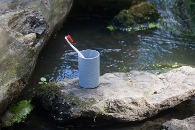 Kırmızı kalkanlı beyaz bir diş fırçası, bir taşın üzerindeki plastik bir bardakta, resim gibi bir nehrin zemininde duruyor. Doğada kendine iyi bakmakla ilgili bir imaj..