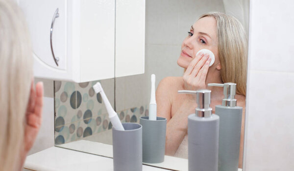 Smiling blonde aged woman in a white towel, stands in the bathroom and takes care of her face, standing by the mirror. Image on the theme of hygiene.
