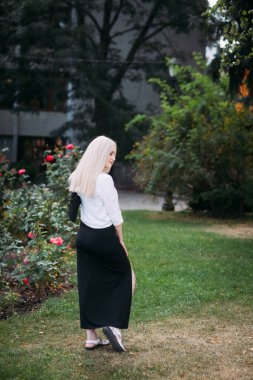 Uzun sarı saçlı, uzun siyah uzun elbiseli ve beyaz gömlekli, güzel, güler yüzlü bir kadın şehirde çiçek açan çalıların arasında parkta yürüyor..