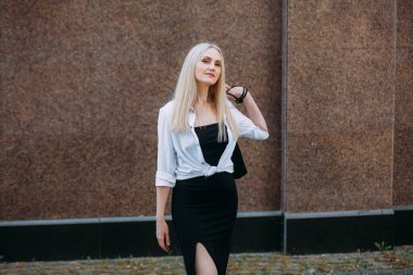 Uzun sarı saçlı, uzun siyah uzun elbiseli ve beyaz gömlekli, güzel, güler yüzlü bir kadın, bir şehir caddesinde duruyor, granit bir bina duvarının arkasında..