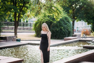 İnce, uzun sarı saçlı, siyah, uzun, seksi elbiseli güzel bir kadın güneşli bir günde şehir parkında çıplak ayakla fıskiyede yürüyor..