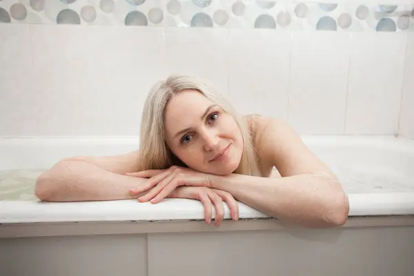 A beautiful smiling girl with blond hair sits in a bath and washes, in the bathroom. An image about cleanliness and about taking care of yourself and your body.