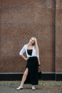 Uzun sarı saçlı, uzun siyah uzun elbiseli ve beyaz gömlekli, güzel, güler yüzlü bir kadın, bir şehir caddesinde duruyor, granit bir bina duvarının arkasında..