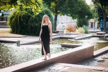 İnce, uzun sarı saçlı, siyah, uzun, seksi elbiseli güzel bir kadın güneşli bir günde şehir parkında çıplak ayakla fıskiyede yürüyor..