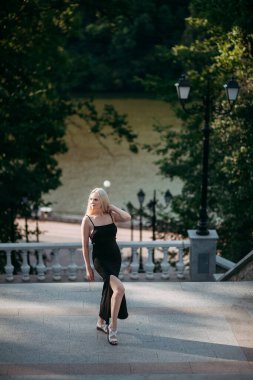 İnce, yaşlı, uzun sarı saçlı, siyah, uzun, seksi elbiseli bir kadın, şehir parkındaki merdivenlerde duruyor. Resimli bir nehrin arka planında..