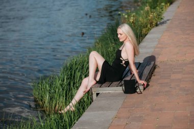 Uzun sarı saçlı, ince bacaklı, siyah uzun elbiseli ve siyah küçük çantalı şık, seksi, yaşlı bir kadın güneşli bir günde, güneşli bir nehrin tepesinde oturur..