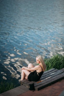 Uzun sarı saçlı, ince bacaklı, siyah uzun elbiseli ve siyah küçük çantalı şık, seksi, yaşlı bir kadın güneşli bir günde, güneşli bir nehrin tepesinde oturur..