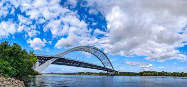 Nehir panoramasının üzerindeki ark köprüsü nehir dalgalarına yansıdı. Bir yaz sabahı şehir manzarası. Dikey detaylı panorama. Yapılarda dalga benzeri çarpıtmalar. Kiev, Ukrayna. Dinyeper Nehri.