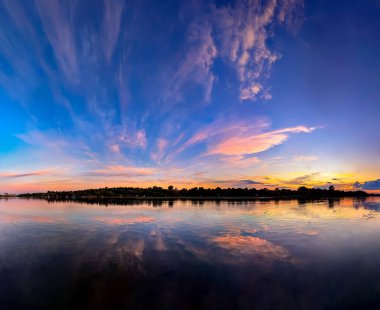 Gün batımı nehre yansıdı. Turuncu ve pembe fan bulutları dağılır. Sakin akşam nehri gökyüzünü yansıtır. Sarı ve turuncu renkler mor gölgelere dönüşür. Su ve bitki örtüsünün geniş panoramik görüntüsü. Temiz su rezervleri. Banyo için su kalitesi.