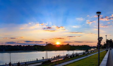 Güneş batıyor, bulutlar mavi ve turuncu dramatik gökyüzünde, güneş ışınlarıyla. Akşam saatlerinin rengini ve ışığını yansıtan nehir kenarında bir gezinti. Sonsuz boşluk ve sükunet