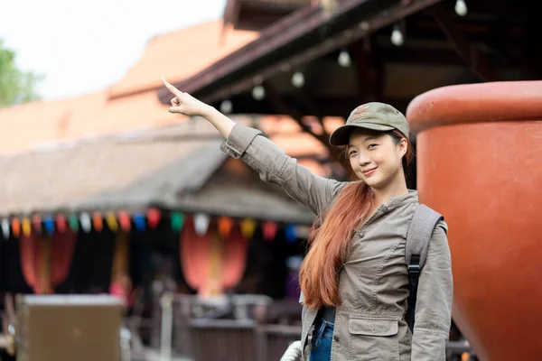 Young asian woman traveler with hat and backpack pointing finger on famous tourist attractions in thailand