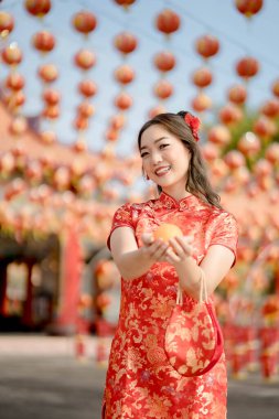 Vertical pictire. Happy Chinese new year. A young lady wearing traditional cheongsam qipao dress holding ancient gold money and bag in Chinese Buddhist temple. Celebrate Chinese lunar new year, festive season holiday. Emotion smile
