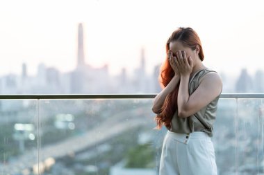 Young asian woman looking away at the city standing on balcony at condo. Frustrated confused female feels unhappy problem in personal life quarrel break up with boyfriend or unexpected pregnancy concept