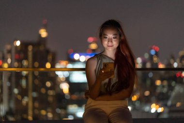 portrait of yong woman using mobile phone while sitting a balcony at condo in the night. Emotion smile