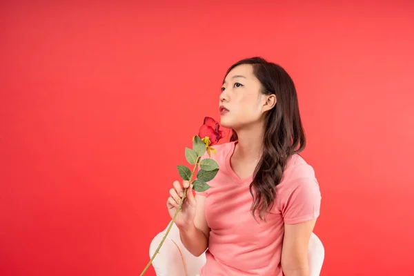 Young Asian woman holding Rose Flower and looking up empty space isolated over red background. Valentines Day concept.