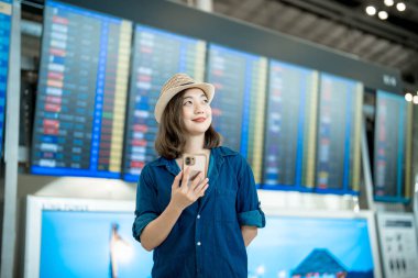 Yolculuğa hazırız. Uluslararası havaalanında cep telefonu kullanan mutlu genç Asyalı kadın uçuş bilgi panosuna bakıyor.