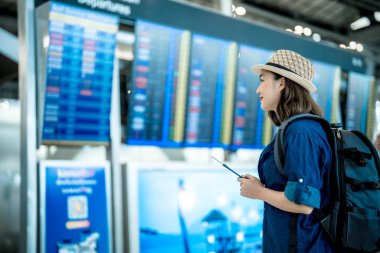 Sırt çantalı genç Asyalı bir kadın uluslararası havaalanında el olarak pasaport ve biniş kartı tutuyor uçuş bilgilerine bakıyor, uçuşunu kontrol ediyor.