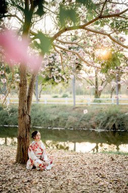 Geleneksel Japon kimonosu giymiş genç bir kadın gün batımında sakin bir nehrin kenarında, kiraz çiçeği ağacının altında meditasyon yapıyor. Huzurlu doğayla çevrili..