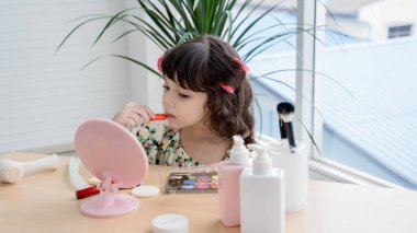 Aynanın önünde makyajla oynayan sevimli küçük bir kız, odaklanarak ve merakla ruj sürüyor, oyuncu bir anda yetişkin güzellik figürlerini taklit ediyor..