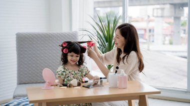 Gülümseyen anne, evinde makyaj seansı sırasında, güzellik aletleri ve kozmetik ürünleriyle eğlenirken, kızına saç bigudileri konusunda yardım ediyor..