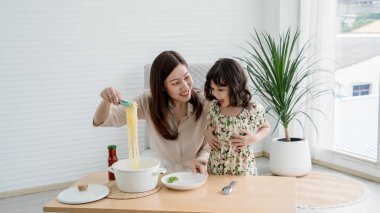 Neşeli anne ve kızı, parlak, modern bir mutfakta makarna pişiriyor, evde yemek hazırlıkları sırasında eğlenceli ve sevgi dolu bir anı paylaşıyorlar..