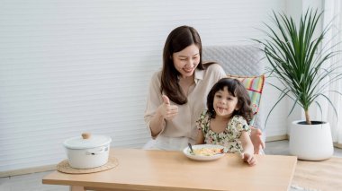 Anne başparmağını kaldırırken, kız yemek masasında spagetti yemekten zevk alıyor. Sıcak, modern bir evde eğlenceli ve lezzetli bir yemek zamanı geçiriyor..