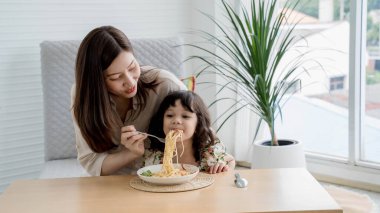 Anne kızını aşk ve özenle spagetti ile besliyor, sıcak bir ev yapımı yemeğin tadını çıkarıyor doğal ışık ve konforlu modern bir yemek alanında..