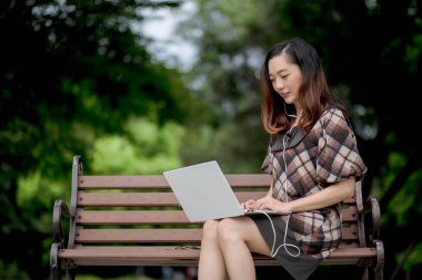Parktaki bankta dizüstü bilgisayar kullanan gülümseyen genç bir kadın, kulaklıkları açık havada ağaçlarla çevrili ve huzurlu yeşil doğa ile eğleniyor..