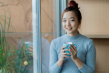 Elinde kahve fincanıyla pencerenin yanında duran gülümseyen Asyalı kadın bardağa yansıyor, doğal ışıkla huzurlu ve rahat bir sabah anının tadını çıkarıyor.
