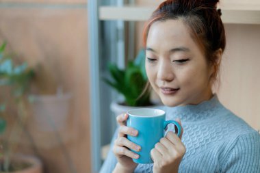 Mavi bir kahve fincanı tutan Asyalı bir kadın, gözleri kapalı, sıcak ve rahat bir mekanda, yumuşak doğal ışıkla içkisinin tadını çıkarıyor.