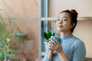 Mavi bir kahve fincanı tutan Asyalı kadın, huzurlu bir memnuniyet içinde gözleri kapalı gülümsüyor, doğal ışık ve bitkilerle sakin ve rahat bir anın tadını çıkarıyor.