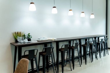 Yüksek ahşap tezgahı, metal bar taburesi, şarap bardakları, çay seti, kolye ışıkları ve en az beyaz arka planı olan modern kafe barı.