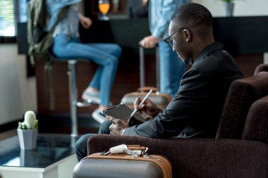 İş adamı, Stylus 'la birlikte dijital tablet kullanıyor. Havaalanı salonunda bekliyor. Etrafı seyahat valizleriyle çevrili ve arka planda sıradan giyinmiş gezginlerle dolu.