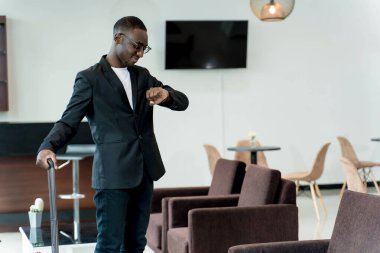 Modern bir havaalanı salonunda kalkış ya da iş seyahatine hazır beklerken saati kontrol eden şık bir işadamı.