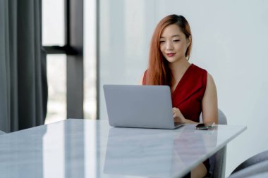 Kendine güvenen Asyalı iş kadını parlak modern ofiste mermer masalı dizüstü bilgisayarda çalışıyor, kırmızı elbise giyiyor ve iş için teknolojiyi kullanırken gülümsüyor.