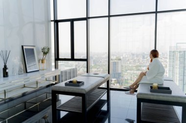 Beyaz cüppeli rahat bir kadın lüks bir kaplıcadan huzurlu bir manzaranın keyfini çıkarıyor. Zarif bir dekor ve doğal gün ışığıyla çevrili masaj yatağında oturuyor.