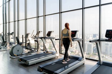 Büyük camlı camları ve panoramik şehir siluetiyle modern yüksek katlı spor salonunda yürüyen bir kadın, sağlık ve zindelik yaşam tarzına odaklanıyor.