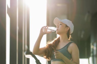Spor salonundan sonra spor salonunda su içen genç bir kadın, güneş ışığında beyaz şapkalı ve spor kıyafetli, hidrasyon ve sağlıklı yaşam tarzını temsil ediyor.