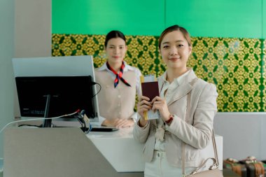 Havaalanı kayıt gişesinde elinde pasaport ve uçuş kartı olan gülümseyen bir kadın. Arka planda havayolu personeli var. Uluslararası seyahat macerasına hazır.