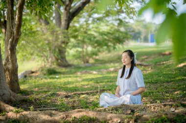 Barışçıl park ortamında yeşil ağaçların altında meditasyon yapan Asyalı kadın, güneş ışığı ve dinginlikle doğal ortamda farkındalık ve iç huzuru uyguluyor.