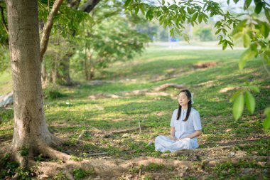 Doğada meditasyon yapan genç bir kadın ağaç gölgesi altında, yeşil parkta huzurlu bir atmosfer, ruhsal ve zihinsel refah için farkındalık ve rahatlama.