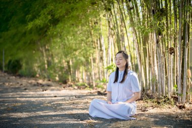 Bambu ormanında huzur içinde meditasyon yapan bir kadın, beyaz geleneksel kıyafetlerle patikada oturuyor, gözleri kapalı, sükunet ve doğal farkındalığın tadını çıkarıyor.