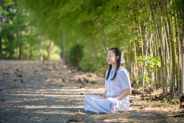 Genç bir kadın, bir orman yolunda, bir bambu koruluğunda huzur içinde meditasyon yapıyor. Huzurlu bir doğal ortamda farkındalık ve ruhani farkındalık uyguluyor.
