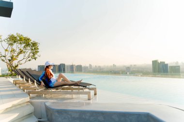 Genç bir kadın, çatı katındaki bir şezlongda panoramik şehir silueti ve arka planda yumuşak bir sabah güneşi olan sonsuz bir havuzun kenarında huzurlu bir anın tadını çıkarıyor.