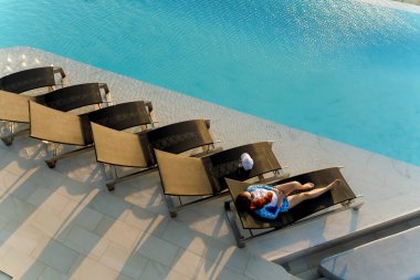 Genç bir kadının güneşlendiği havadan bir şezlongda tek başına çatı katındaki sonsuz havuzun kenarında köpüklü suyla yaz güneşinin ve huzurlu anın tadını çıkardığı görüntüsü.