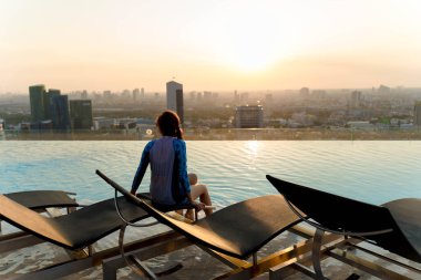 Gün batımında çatıda sonsuz bir havuz kenarında oturan kadın, huzurlu şehir manzarasının keyfini çıkarıyor. Sıcak güneş ışığı sakin bir şehir ortamında suyu yansıtıyor.