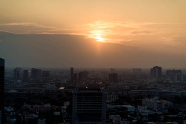 Güneşin kısmen bulutların arkasına gizlendiği dramatik şehir silueti kentsel binalar ve kasvetli atmosferik ufuklar üzerine sıcak turuncu ışık saçıyor.
