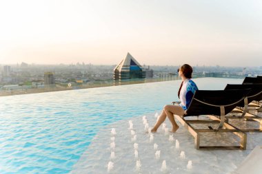 Çatı katındaki sonsuz havuzda dinlenen bir kadın, ayak çeşmeleri, panoramik şehir manzarası ve huzurlu şehir kaçışında yumuşak güneş ışığının tadını çıkarıyor.